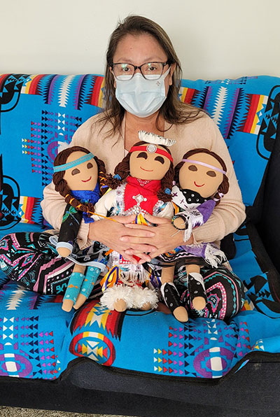 Elaine McArthur sitting on a sofa, wearing a mask, and holding three dolls that are dressed in traditional regalia