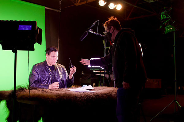 Lancelot Knight sits behind a news desk with Muskwa Lerat standing next to him