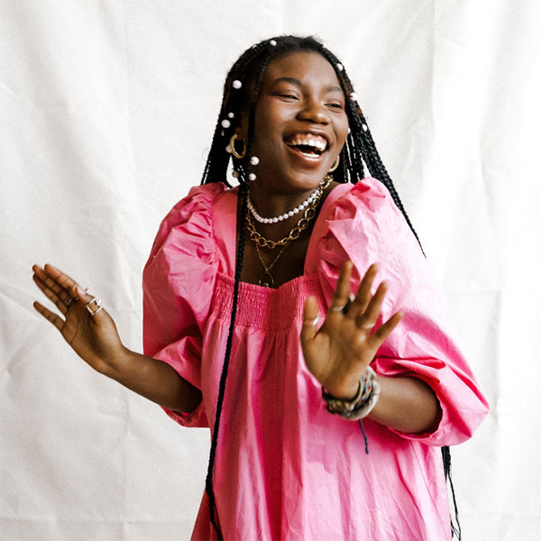 Peace Akintade, black young woman in a pink dress and braids, with a big smile touching her forehead and holding up both hands in candid pose