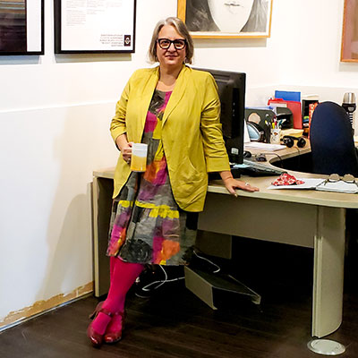 Belinda Harrow, SK Arts Permanent Collection Consultant - Older woman leaning on a desk with crossed legs holding a coffee mug. She has grey hair, wearing glasses and smiling. She is wearing a mustard yellow jacket, a colourful dress, red stockings and strapped shoes.