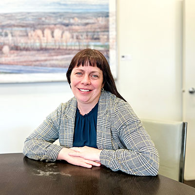Jody Greenman-Barber, Program Consultant - White woman in an office. She has brown hair, bangs, is wearing a plaid jacket and black blouse. There is a painting behind her.