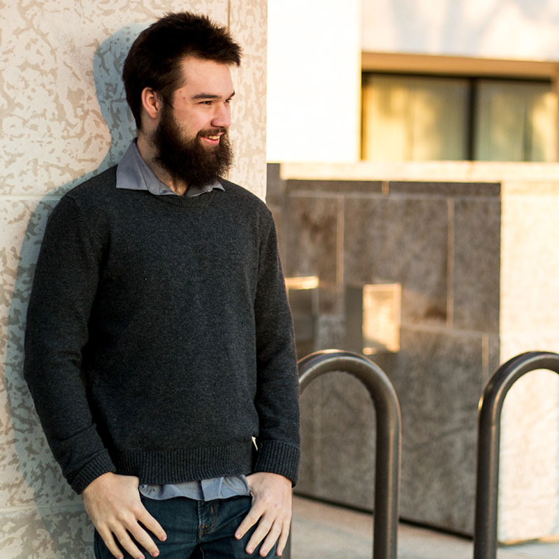 John Loeppky - White male leaning against a wall. He as short hair, a beard and wearing a plaid collar shirt with a black sweater over it and jeans, His thumbs are hooked in his pockets.
