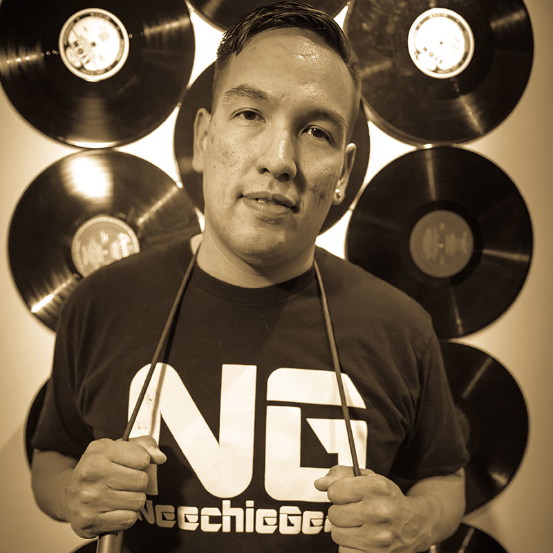 Bradley Bellegarde - Portrait of Indigenous man standing in front of records on a wall. He is holding a string on his neck.