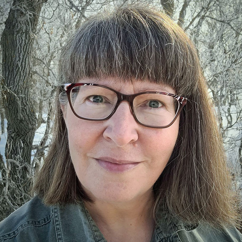 Brenda Schmidt - Indigenous woman with short grey hair. She is wearing bangs and glasses.