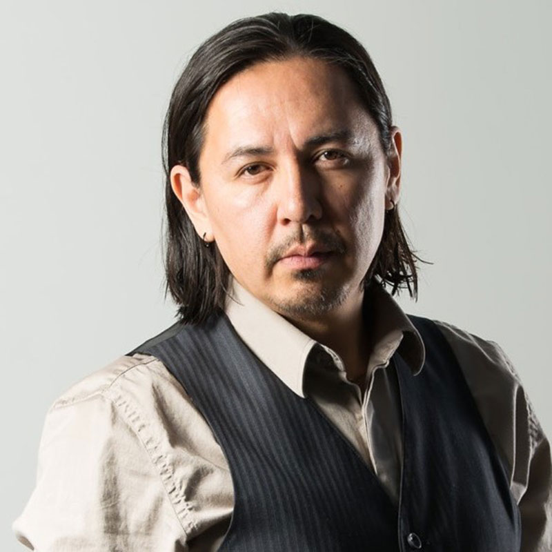 Curtis Peeteetuce - Headshot of indigenous man wearing a cream and black three-piece suit with a bow tie and suit without the jacket. His medium length hair is combed back.