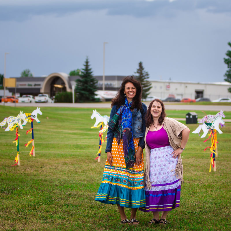 Danielle Castle - Two women standing in a field smiling with artwork in the shape of horses sticking up out of the ground