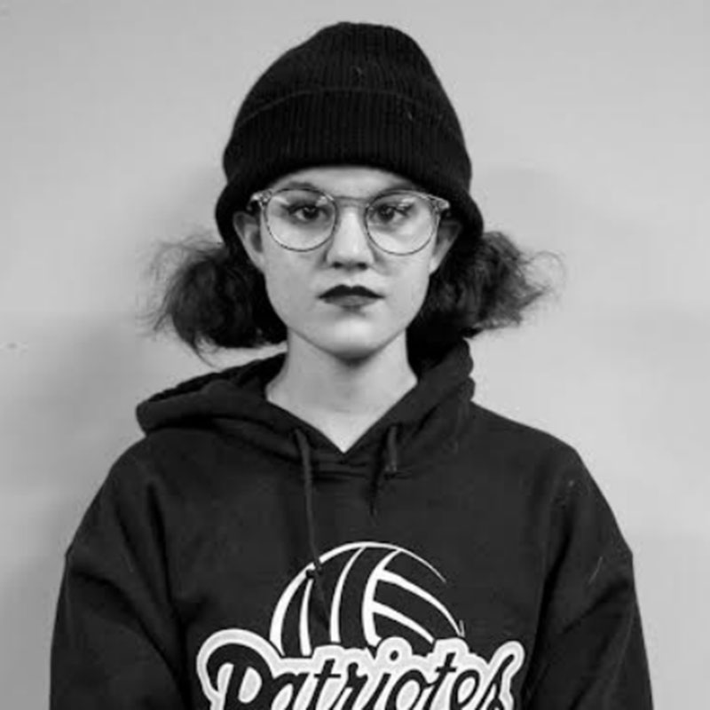 Kam Miller - Young girl in black and white photo wearing a beanie, glasses, a hoodie and lipstick. Her hair is in buns.