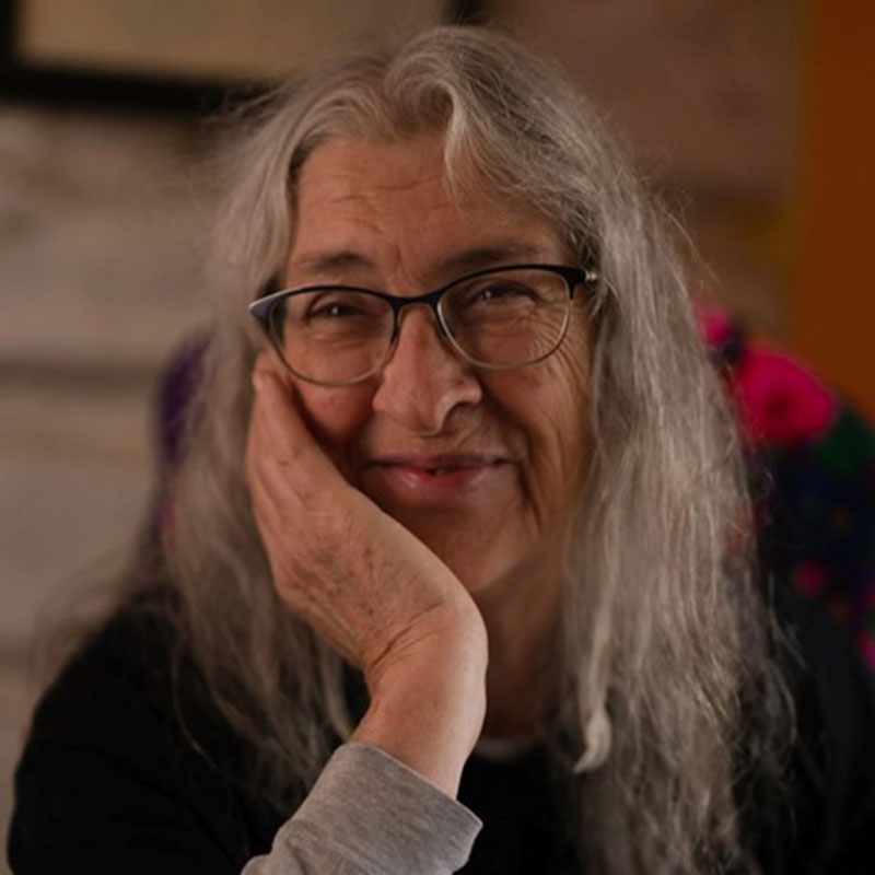 Marjorie Beaucage - Métis woman with long grey hair wearing glasses and smiling with one hand on her cheek