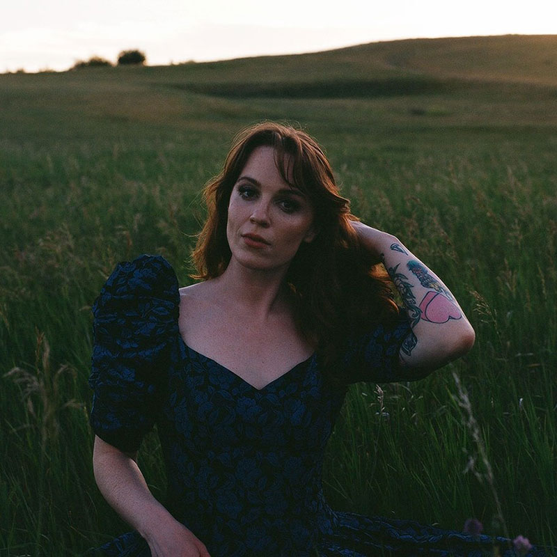 Megan Nash - A women in a field with red hair, she is wearing a blue dress with big sleeves and had one arm covered in tattoos going through her hair