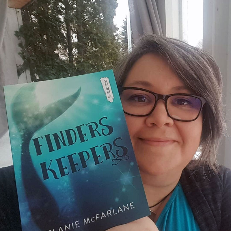 Woman, Melanie McFarlane wearing glasses, smiling and holing a book, titled Finders Keepers next to her face