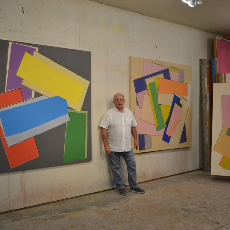 Robert Christie - Man standing in art studio between two large art pieced hung on a wall and other stacked in a corner