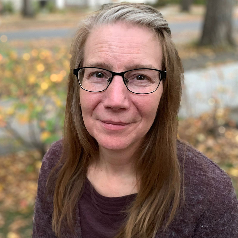Theressa Slind - Close up portrait of woman. She has long grey and brown hair. She is smiling, wearing glasses and wearing a purplish brownish sweater.