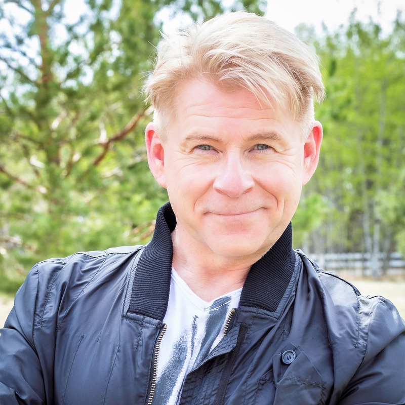 Anthony Bidulka - Older man wearing a navy blue jacket and white t-shirt smiling. There are green trees in the background.