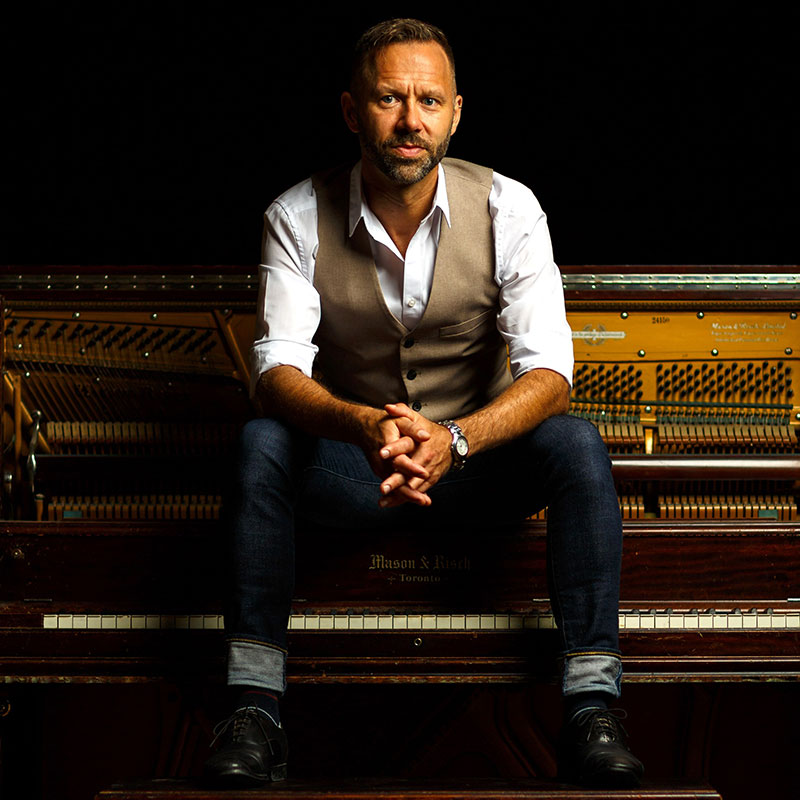 Jeffery Straker - man sitting on a vintage piano wearing a white shirt, brown vest, blue jeans and black boots