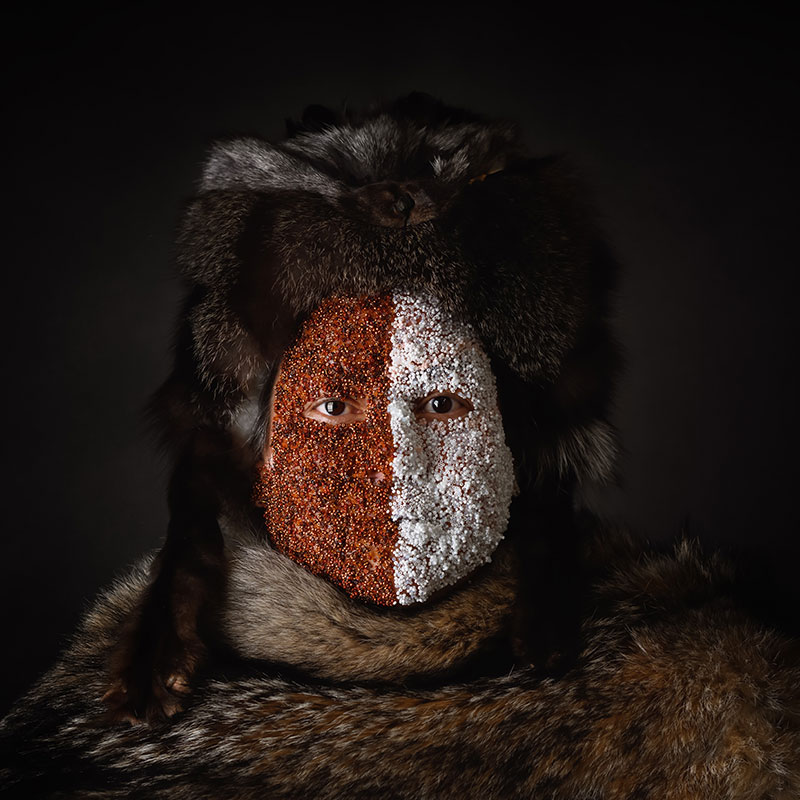 Marcy Friesen - Half Breed - Photo of Indigenous woman wearing fur hat and cloak and white beads on half of her face and brown beads on the other half