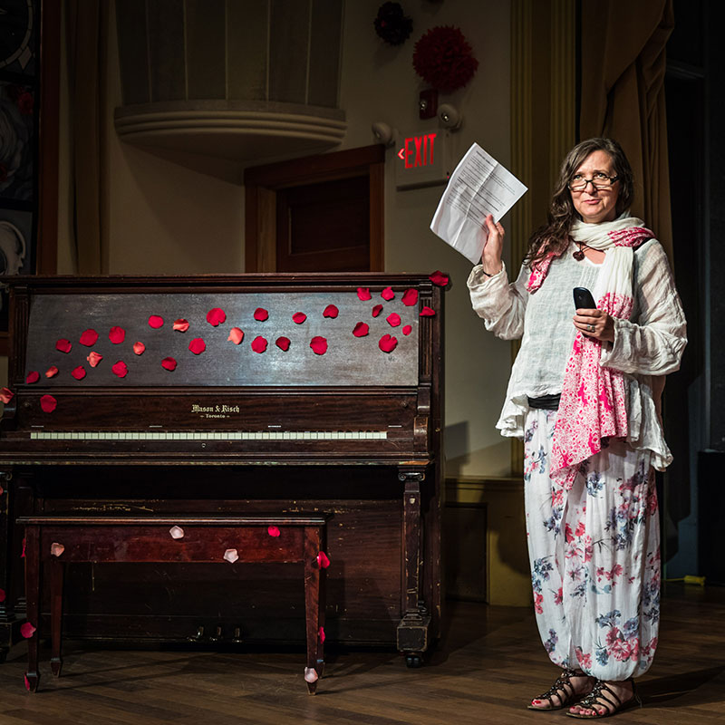 Traci Foster - Woman wearing glasses on stage wearing a long sleeve top, a long skirt and a scarf. She is holding up a sheet of paper and standing next to a piano.