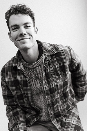 Andrew Robertson - Black and white portrait of Canadian Artist from Shaunavon Saskatchewan. He is wearing a t-shirt under a plaid shirt, leaning forward with one hand behind him and has a closed mouth smile.