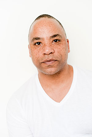 Chancz Perry joins SK Arts- Headshot of black man with freckles wearing a white v neck t-shirt and a white background.
