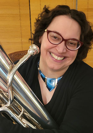 Amanda McArthur for SK Arts Arts of Celebration 2025. Photo of smiling woman wearing red glasses and holding a tuba.