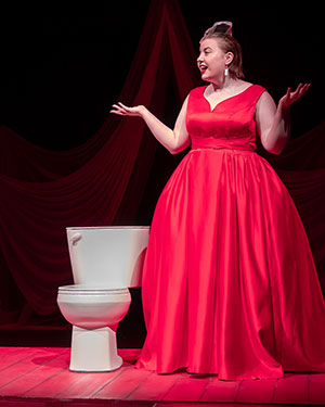 Avianna Hudym by Ash for SK Arts Arts of Celebration 2025. Woman wearing a red gown on stage in that theatre place with a toilet on set.