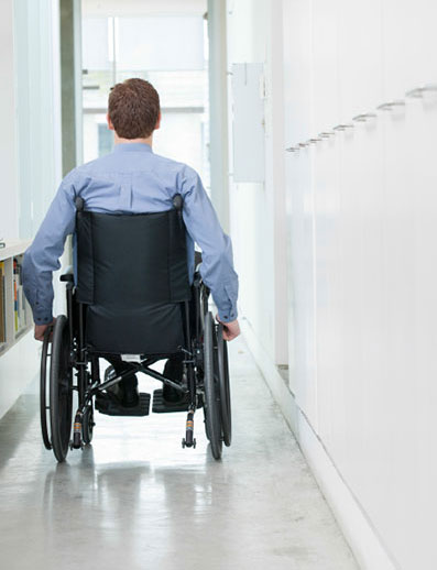 Picture of rear view of white presenting male wearing a powder blue shirt in a wheelchair moving down a white hallway
